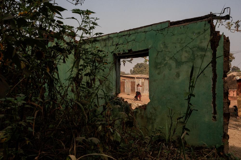 A woman and her child are seen through the window of a destroyed house near the PK5 neighbourhood in Bangui. The town and surrounding area have been the scene of often brutal violence for the last 10 years. Central African Republic, January 2023. © Adrienne Surprenant/MYOP