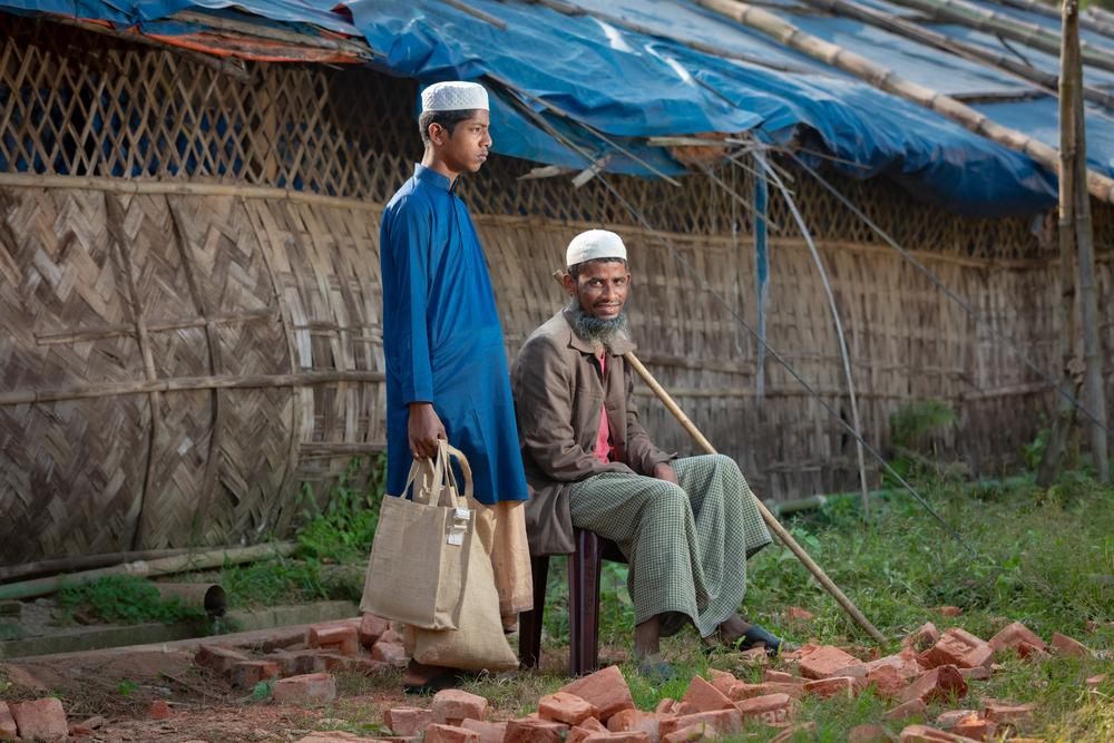 Abdulshakour sits while his son stands beside him, carrying the bags that hold the few belongings his family managed to bring from home while fleeing the attacks on Rohingya people in Myanmar in August 2017. Abdulshakour now lives with his family, including his seven children, in the Cox’s Bazar refugee camp. Bangladesh, December 2022. © Mahammad Hijazi/MSF