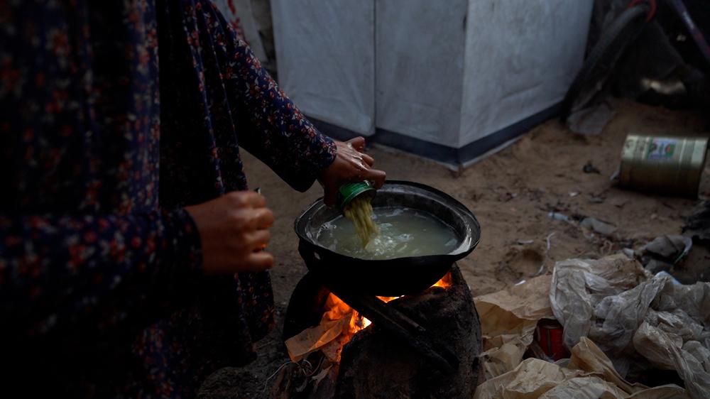 A mother prepares a meal for her family and relatives, pouring a can of green beans into boiling water over a fire she lights herself.